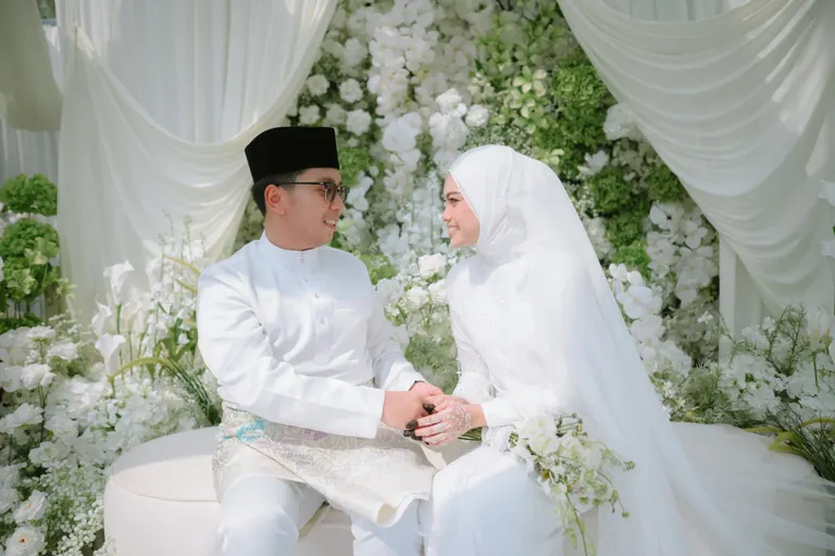 A couple seated hand in hand at their nikah, surrounded by white flowers